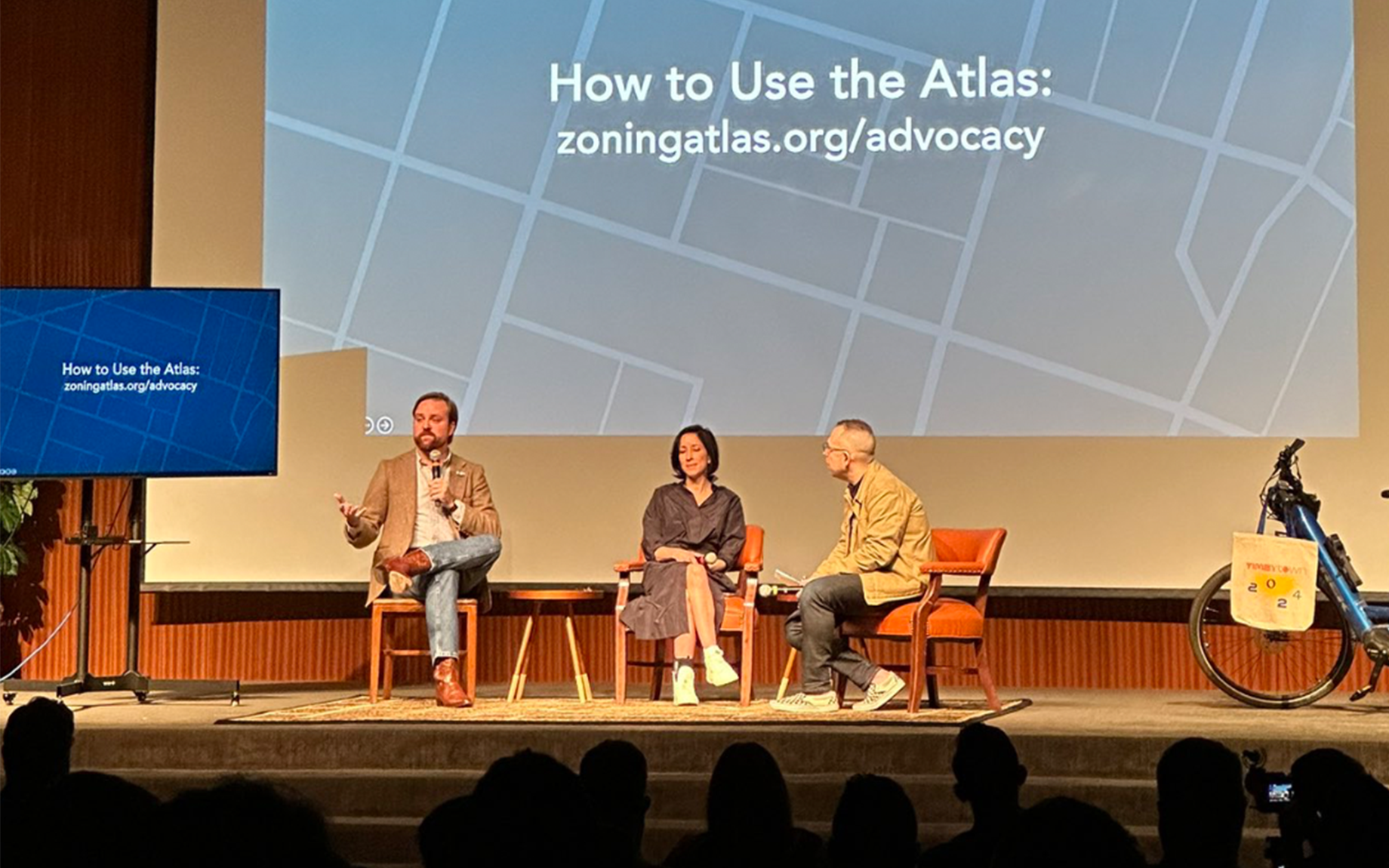 Three people sit on stage with a screen which reads, "How to Use the Atlas"