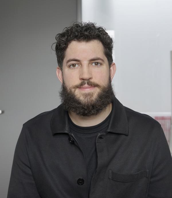 A man with dark curly hair and a dark beard looking at the camera in front of a grey wall